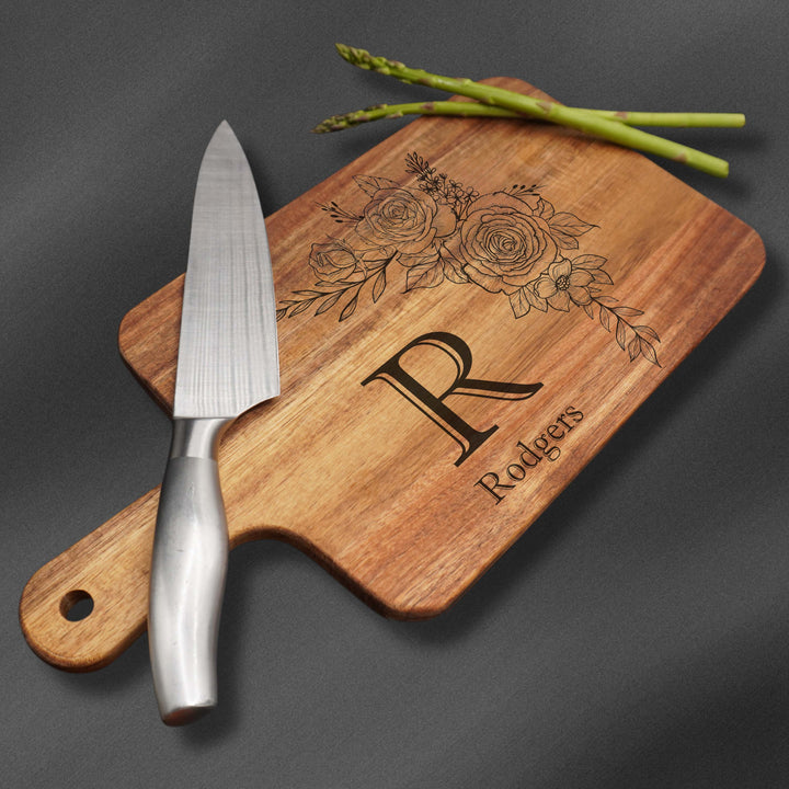Cutting board with handle and an initial with a last name and flower with a knife and asparagus. 